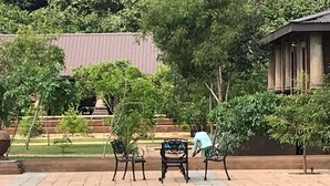 Un espacio al aire libre con sillas y mesas, rodeado de árboles y un edificio con columnas.