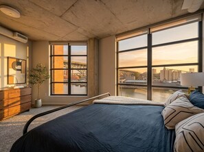 Ein Schlafzimmer mit einem großen Bett, Blick auf eine Stadtkulisse durch große Fenster, einer Topfpflanze und einer Holzkommode.