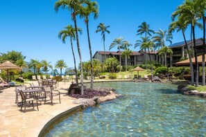 A clear pool surrounded by palm trees and lounge chairs, with a building in the background. - Kauai Escape at Award-Winning Bay Resort | Outdoor Swimming Pool | Parking (Princeville)