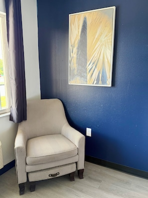 Habitación de esquina con un sillón estampado, pared azul y una obra de arte enmarcada.