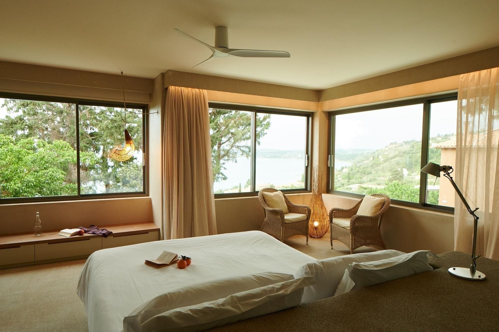A hotel room with a bed, two wicker chairs, a table, and large windows offering a view of the outdoors.