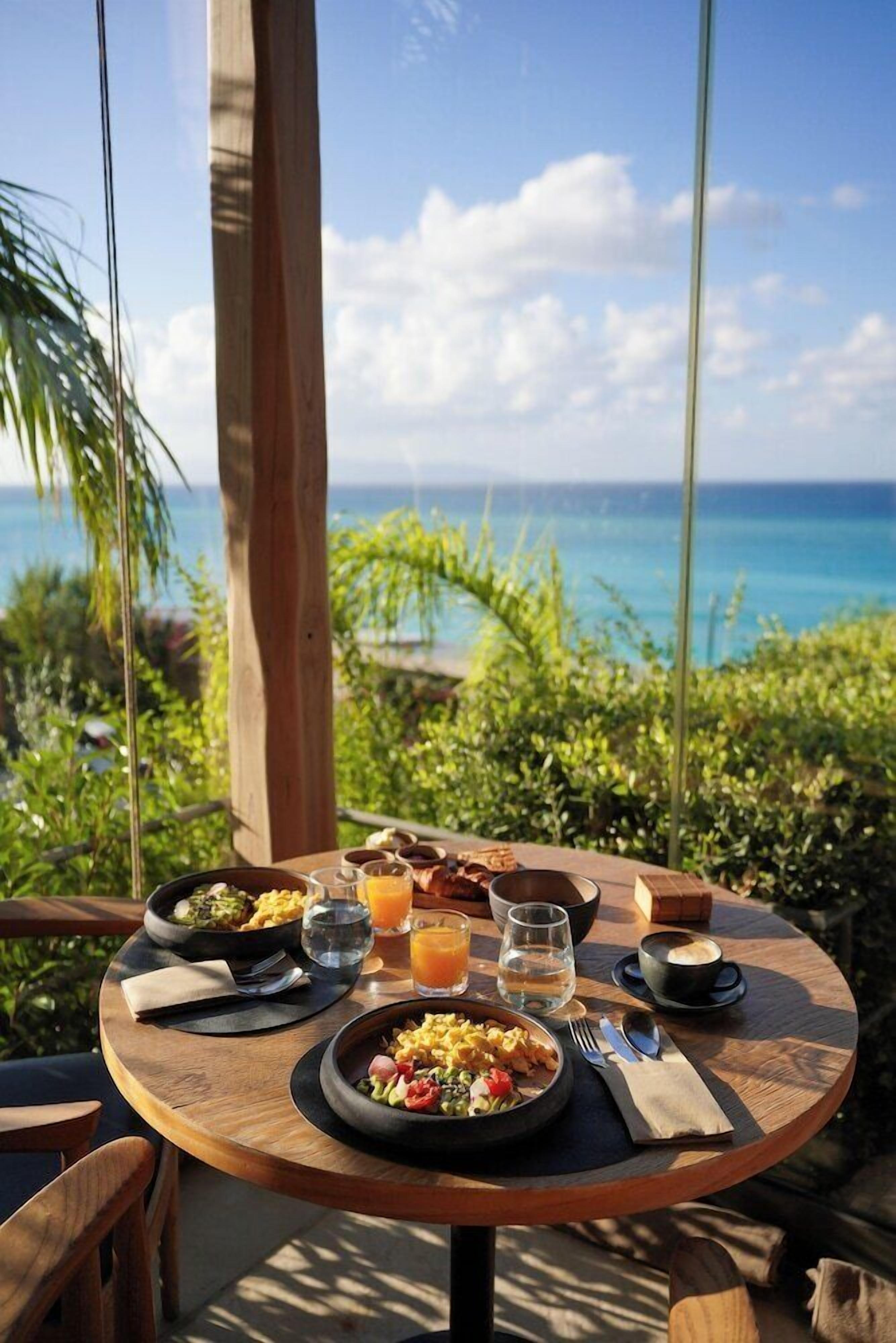 A round wooden table with breakfast items, including eggs, toast, and fruit, set against a backdrop of a clear blue sky and ocean view.