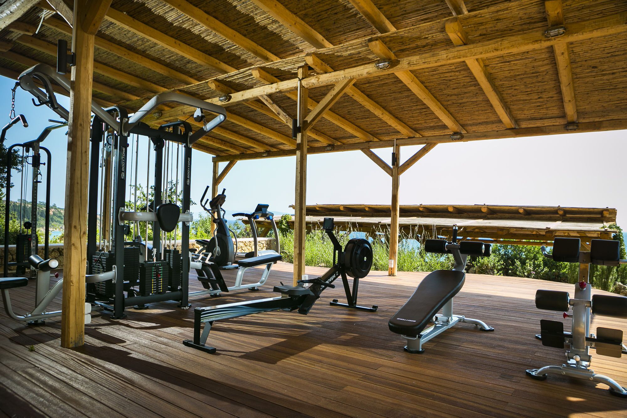 An outdoor gym with various exercise equipment, including a bench, weight machines, and cardio machines, under a wooden structure.