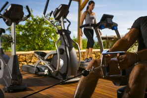 A gym with stationary bikes and a treadmill, a person using a stationary bike, and another person walking on a treadmill. - F Zeen (Kefalonia)