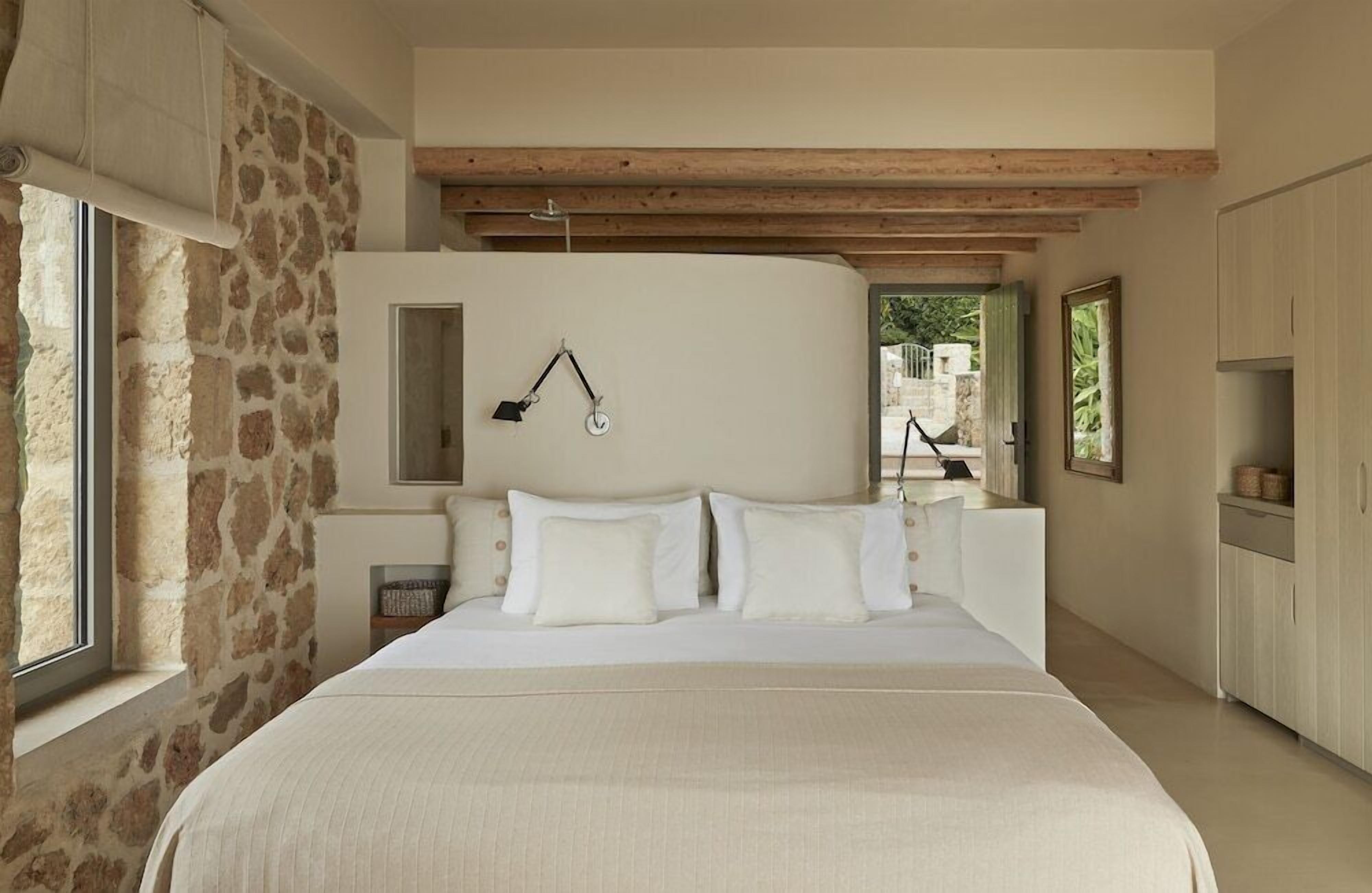 A bedroom with a large bed, stone walls, wooden beams, and a view through an open door.