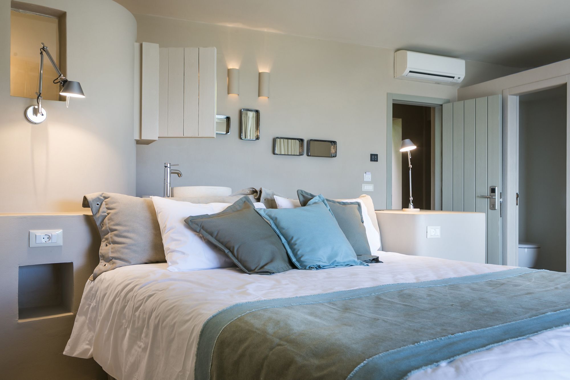 A bedroom with a bed, bedside lamps, a mirror, a wall-mounted light, and an air conditioning unit.