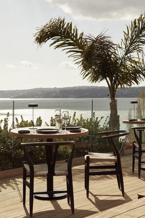 An outdoor dining area with tables and chairs, overlooking a body of water. - F Zeen (Kefalonia)