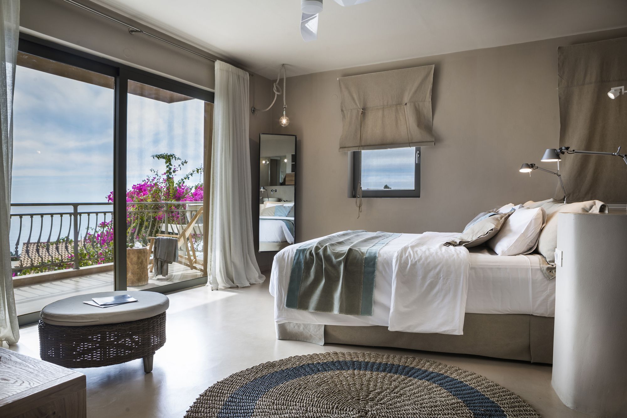 A modern bedroom with a large bed, a round side table, a view of the sea through a balcony, and a mounted TV.