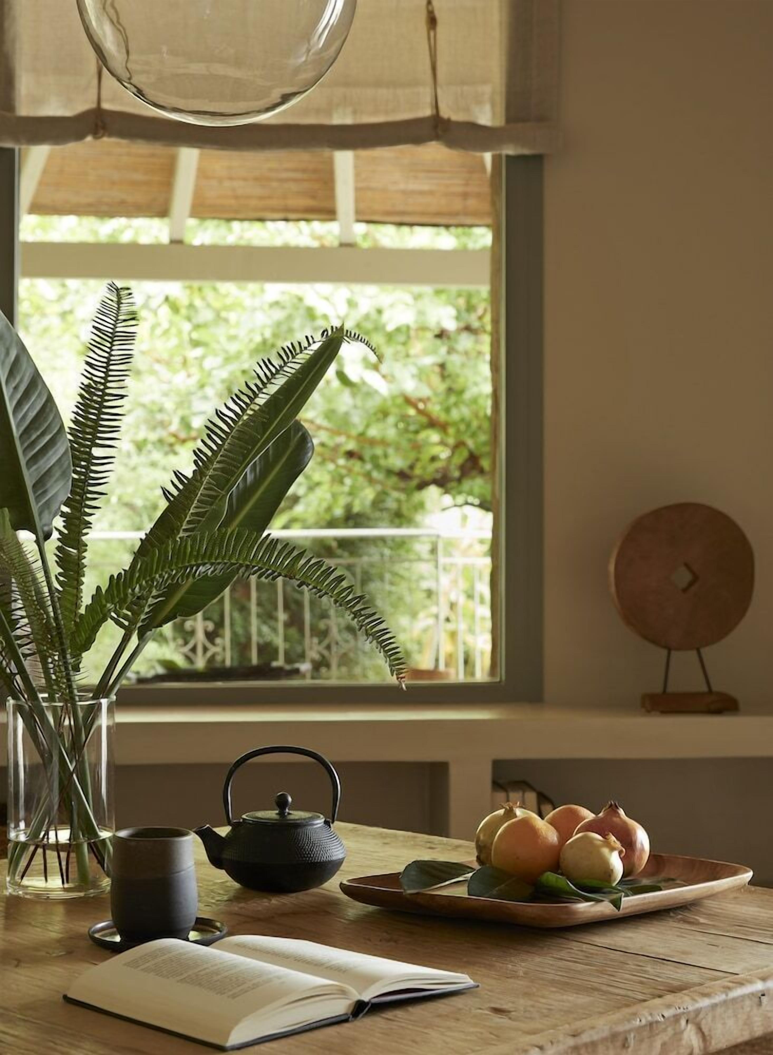 A room with a window, a potted plant, a teapot, a tray with fruits, and a book on a table.