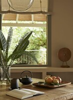 Una habitación con ventana, una planta en maceta, una tetera, una bandeja con frutas y un libro sobre una mesa.