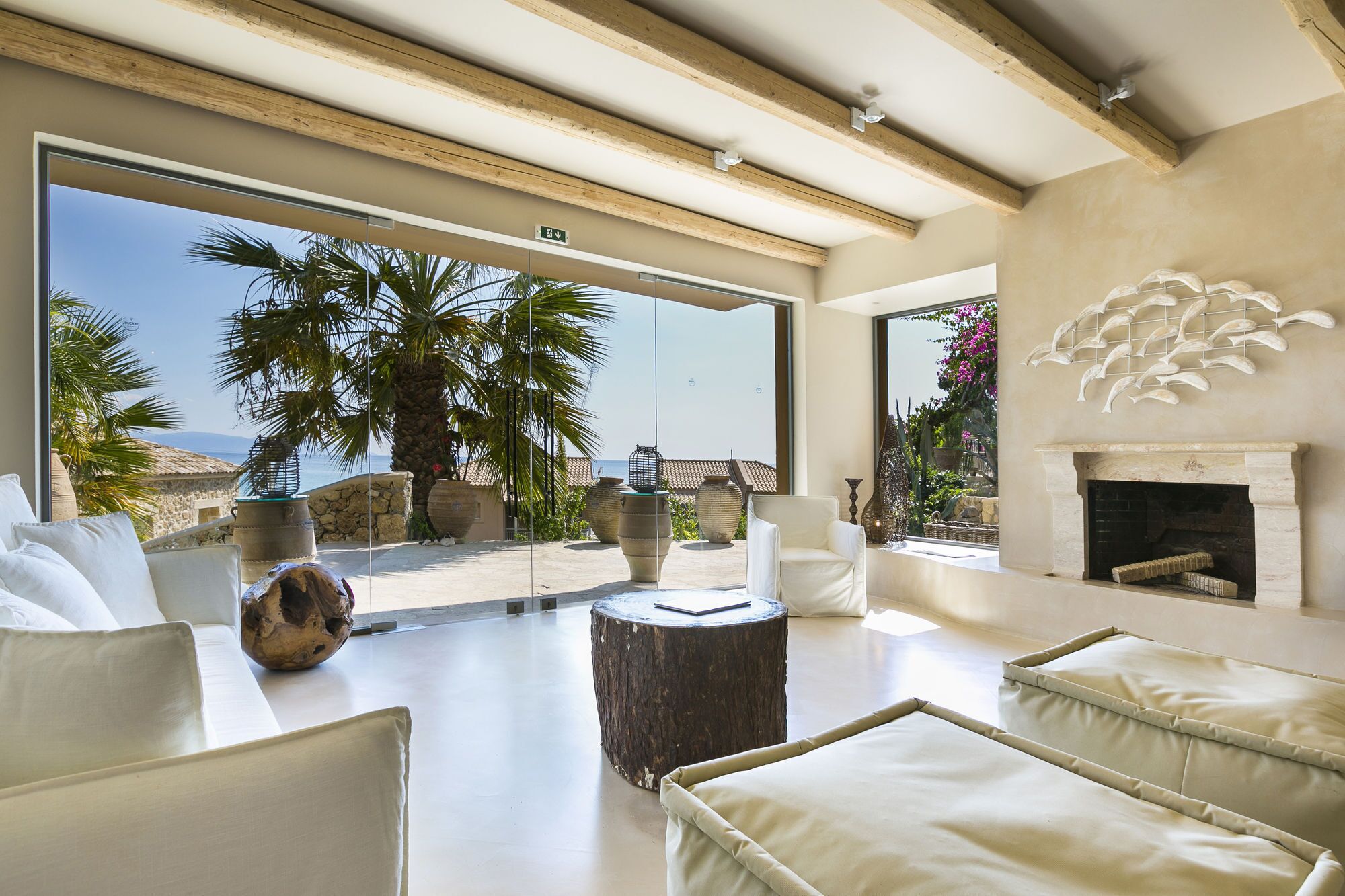A spacious living room with a large glass door leading to a balcony, a fireplace, and a view of palm trees and the sea.