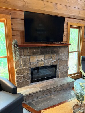 A stone fireplace with a flat-screen TV mounted above it, flanked by wooden walls and large windows. - Pet-Friendly Cabin Near Pigeon Forge & Gatlinburg (Pigeon Forge)