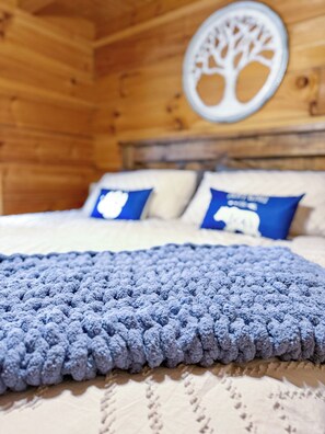 Ein Bett mit einer blauen Strickdecke, weißen Kissen und einem blauen Kopfkissen mit einem Bär-Motiv.
