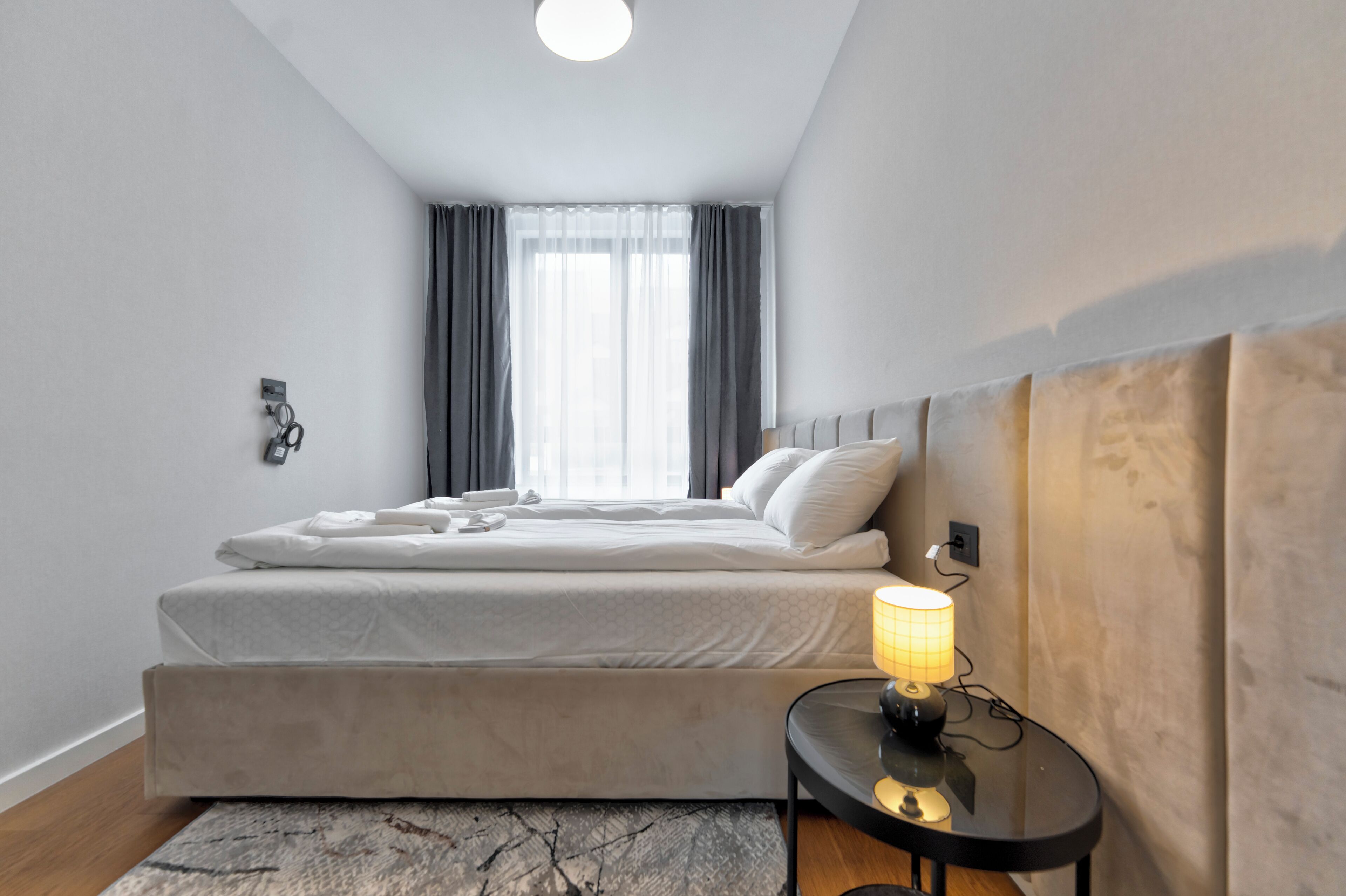 Une chambre d’hôtel moderne équipée d’un lit, d’une table de chevet avec une lampe, d’une fenêtre avec des rideaux et d’un téléphone fixé au mur.