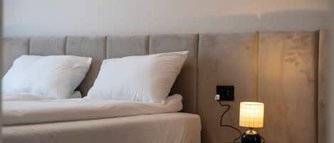Una cama bien hecha con sábanas y almohadones blancos, una mesita de noche con lámpara, e un enchufe de pared.