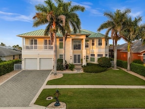 A two-story house with a green roof, a front garage, and a well-maintained lawn with palm trees.