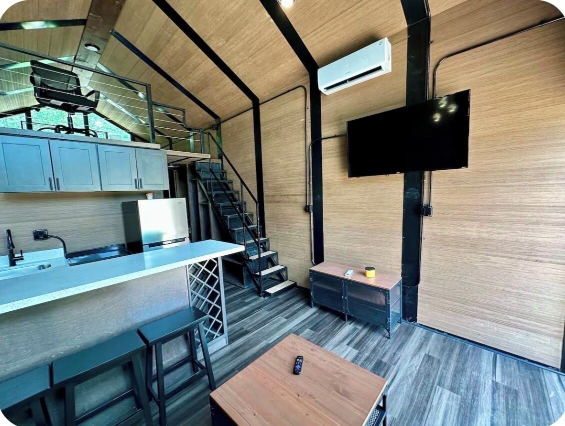 A modern kitchen with a built-in stove, a dining area with a table and chairs, a staircase, and a mounted TV.