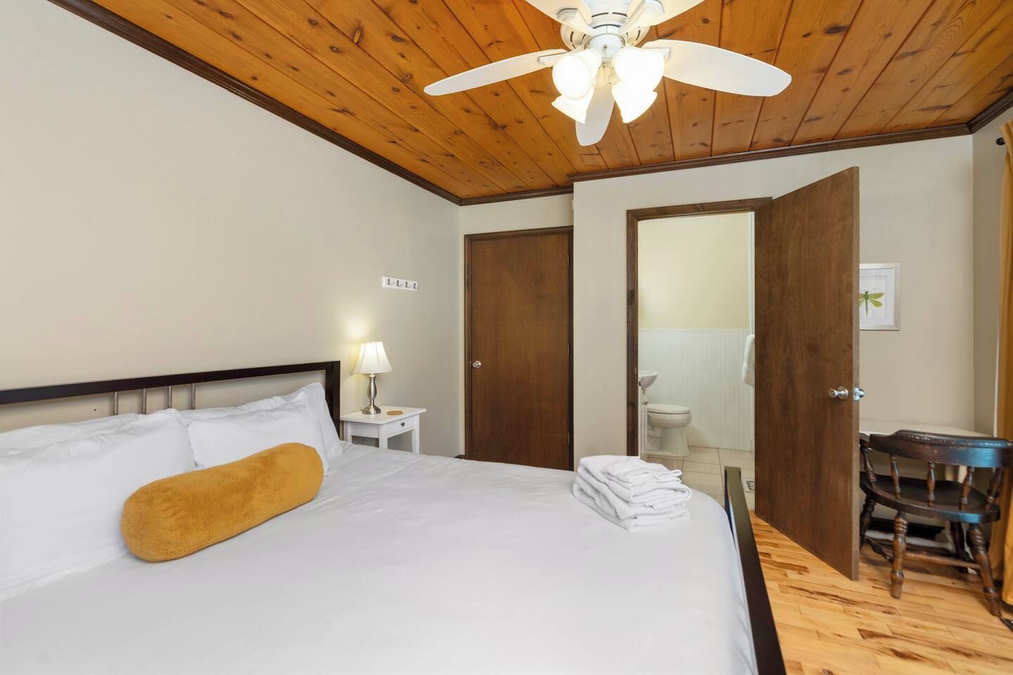 Habitación de hotel con techo de madera, cama con ropa blanca y una sola almohada marrón, mesita de noche con lámpara, una silla y puerta que da a un baño.