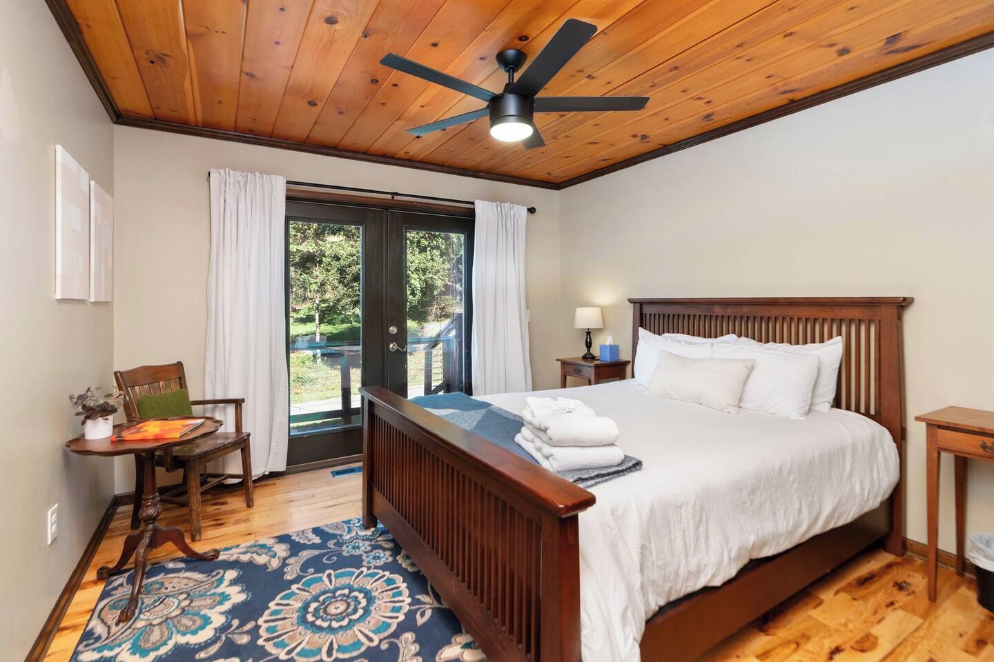 Une chambre avec un plafond en bois, un lit recouvert de draps blancs, une table de chevet, une chaise et une vue sur l’extérieur, visible à travers une porte vitrée.