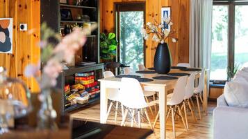 A wooden interior with a dining table, chairs, a vase with flowers, and a shelf with board games.