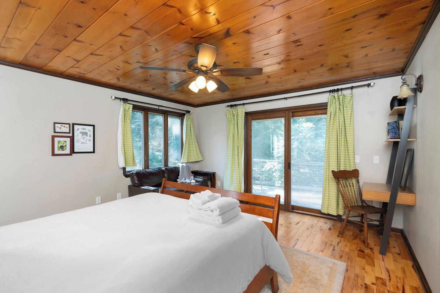 Ein Schlafzimmer mit einem Bett, einem Schreibtisch, einem Stuhl, einem Deckenventilator und großen Fenstern mit Vorhängen.