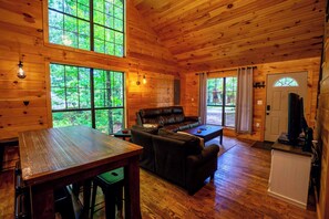 Interior de una cabaña con techo de madera, ventanas amplias, un sofá de cuero, una mesa de comedor y una pequeña cocina.