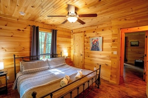 Dormitorio de cabaña de madera con cama, ventilador de techo, un cuadro y una puerta que da a otra habitación.