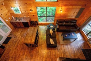A cabin interior with a wooden floor, a dining table, a sofa, a coffee table, and a large window with a forest view. - Rosebud | Hot Tub | WiFi | 2 Min Gorge Underground | 6 Min to Natural Bridge State Park (Gillmore)