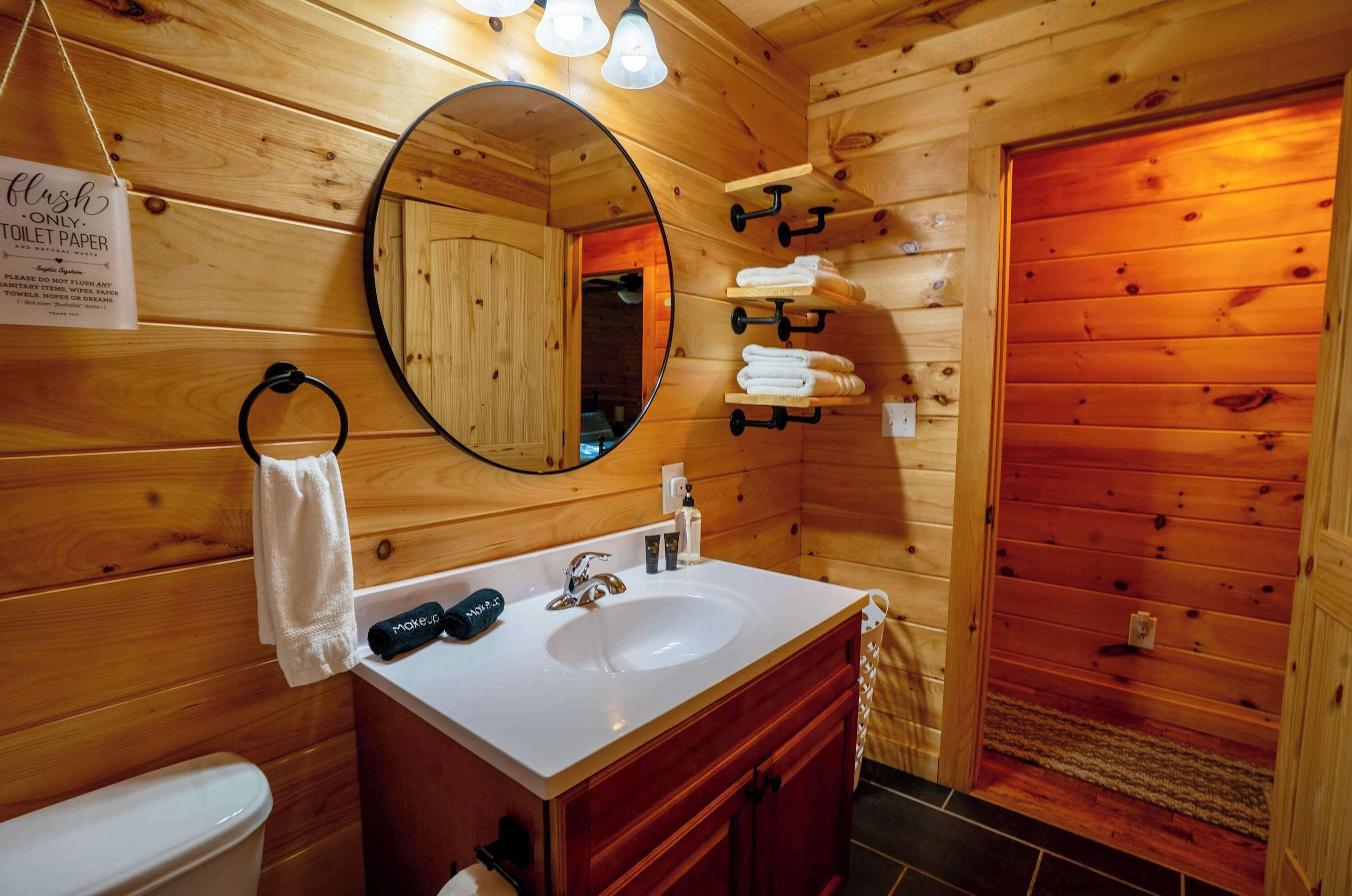 A wooden cabin bathroom with a sink, mirror, towel rack, and a sign indicating toilet paper usage.