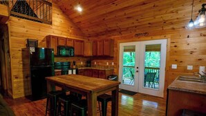 Interior de uma cabana de madeira com cozinha, sala de jantar e varanda com vista para a floresta.
