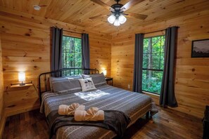 Un dormitorio de cabaña de madera con una cama, mesitas de noche, un ventilador de techo y ventanas con cortinas.