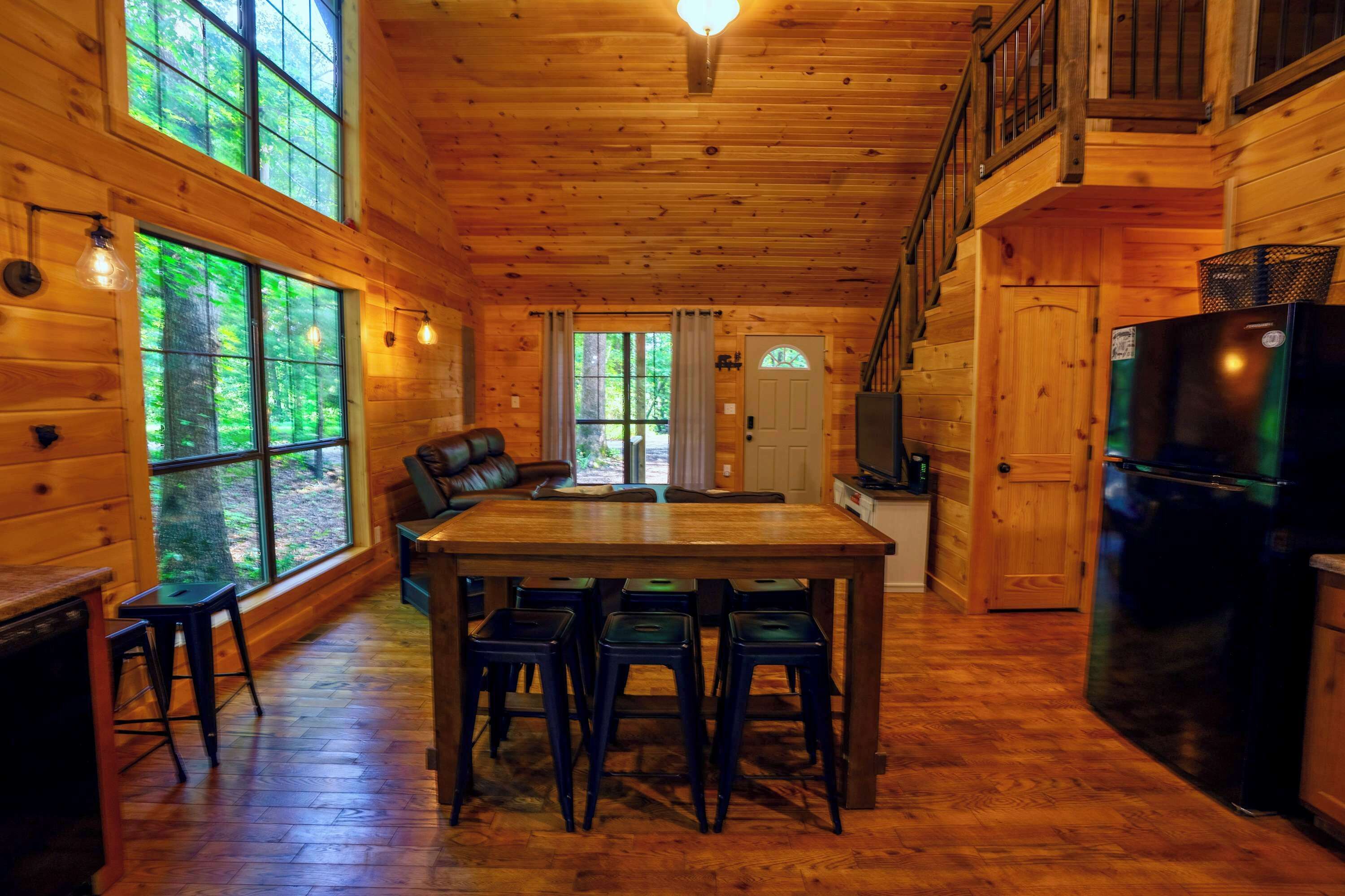 A wooden cabin interior with a dining area, kitchen, and large windows.