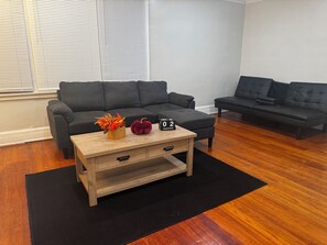 A modern living room with a dark grey sofa, a wooden coffee table with a basket and decorative items, and a black rug on a wooden floor. - Adorable 2-bedroom apartment with AC in cool Detroit (Detroit)