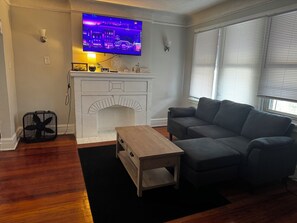 Een woonkamer met een open haard, een flatscreen televisie erboven en een grijze bank met een houten salontafel.
