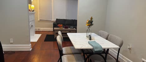 A dining area with a white table, chairs, and a vase with flowers. There is a sofa and a window with blinds in the background.