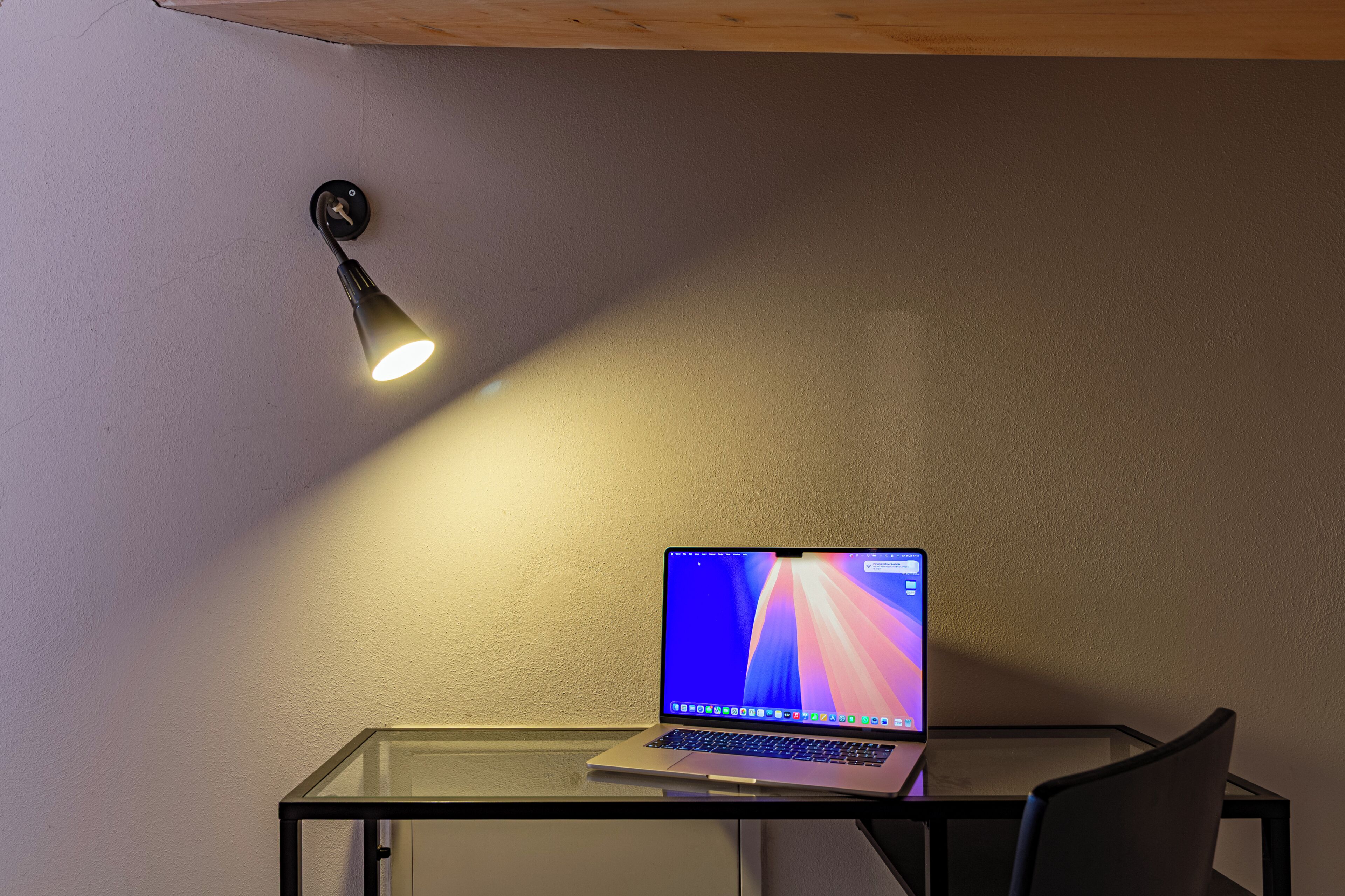 A laptop on a glass table with a wall-mounted lamp above it.