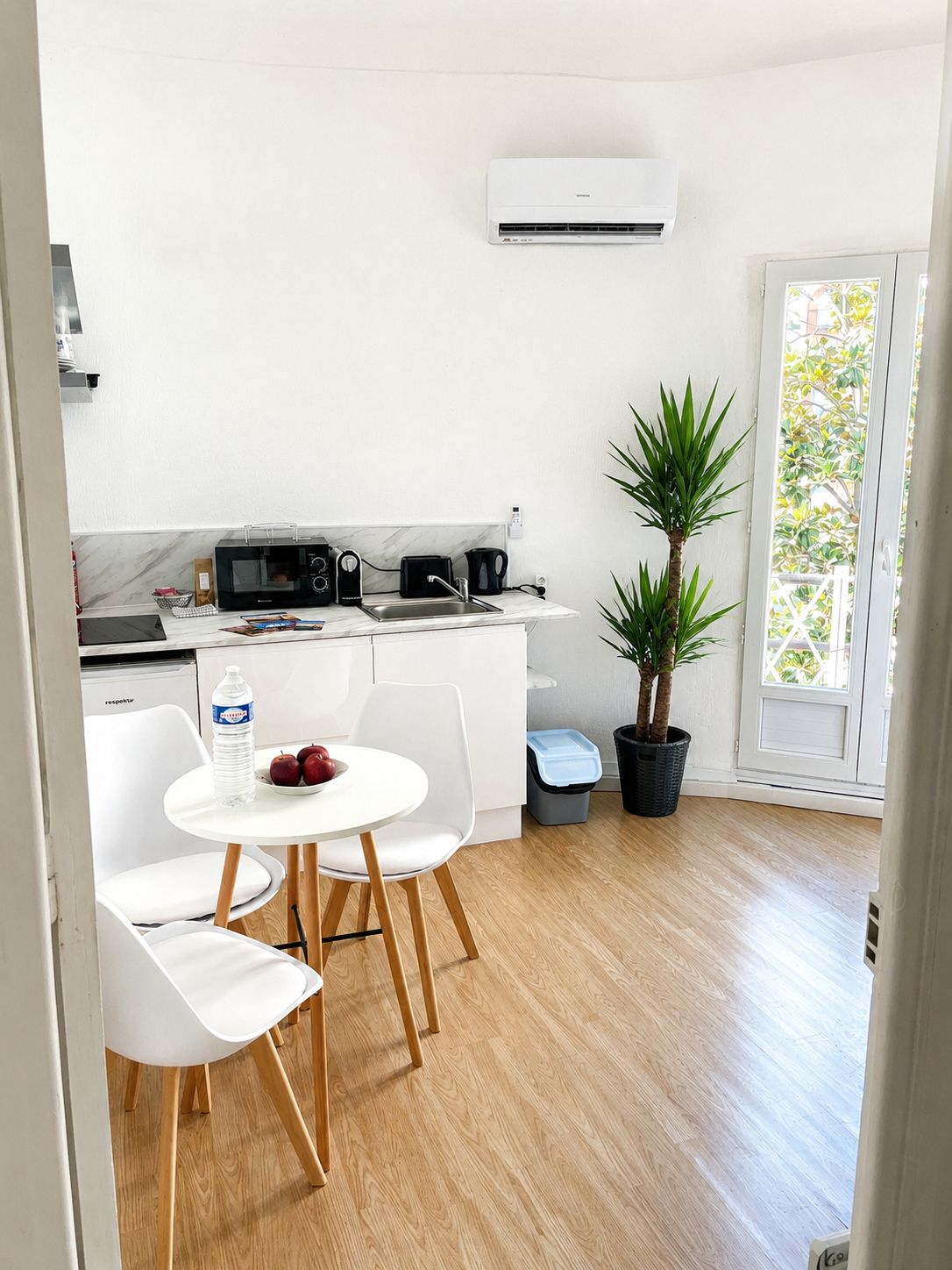 Cocina moderna con armarios blancos, microondas, cafetera y un lavamanos. Hay un comedor con mesa y sillas blancas, y una planta en maceta.