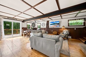 Un salon spacieux doté d’un plancher en bois, d’un canapé gris, d’une table à manger avec des chaises, d’un téléviseur fixé au mur et d’une grande fenêtre avec des rideaux.