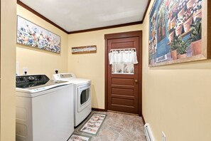 Een wasserette met een wasmachine en droger, een deur met gordijn, en een muur met een bloemschilderij.