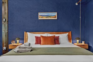 A bedroom with a blue wall, a wooden headboard, a bed with white bedding, red and orange pillows, a green throw blanket, and a framed landscape picture above the bed.