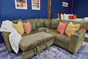 A green sectional sofa with a white throw blanket, red and yellow patterned pillows, and a blue and white patterned rug.
