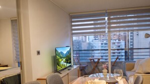 A dining area with a wooden table set for two, a television displaying a landscape, and a view of a cityscape through large windows. - Santiago Barrio Italia - Domus Travels (Santiago)