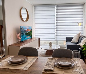 Ein Essbereich mit Tisch für zwei Personen, einer Couch, einem Fernseher und einem Fenster mit Jalousien.