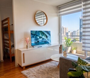 Ein modernes Wohnzimmer mit Fernseher, einer Holzleiter, einem Blumentopf und Blick auf die Stadt durch große Fenster.