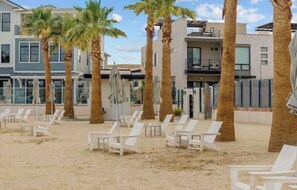 A beachfront property with multiple-story buildings, palm trees, and white lounge chairs on the sand.