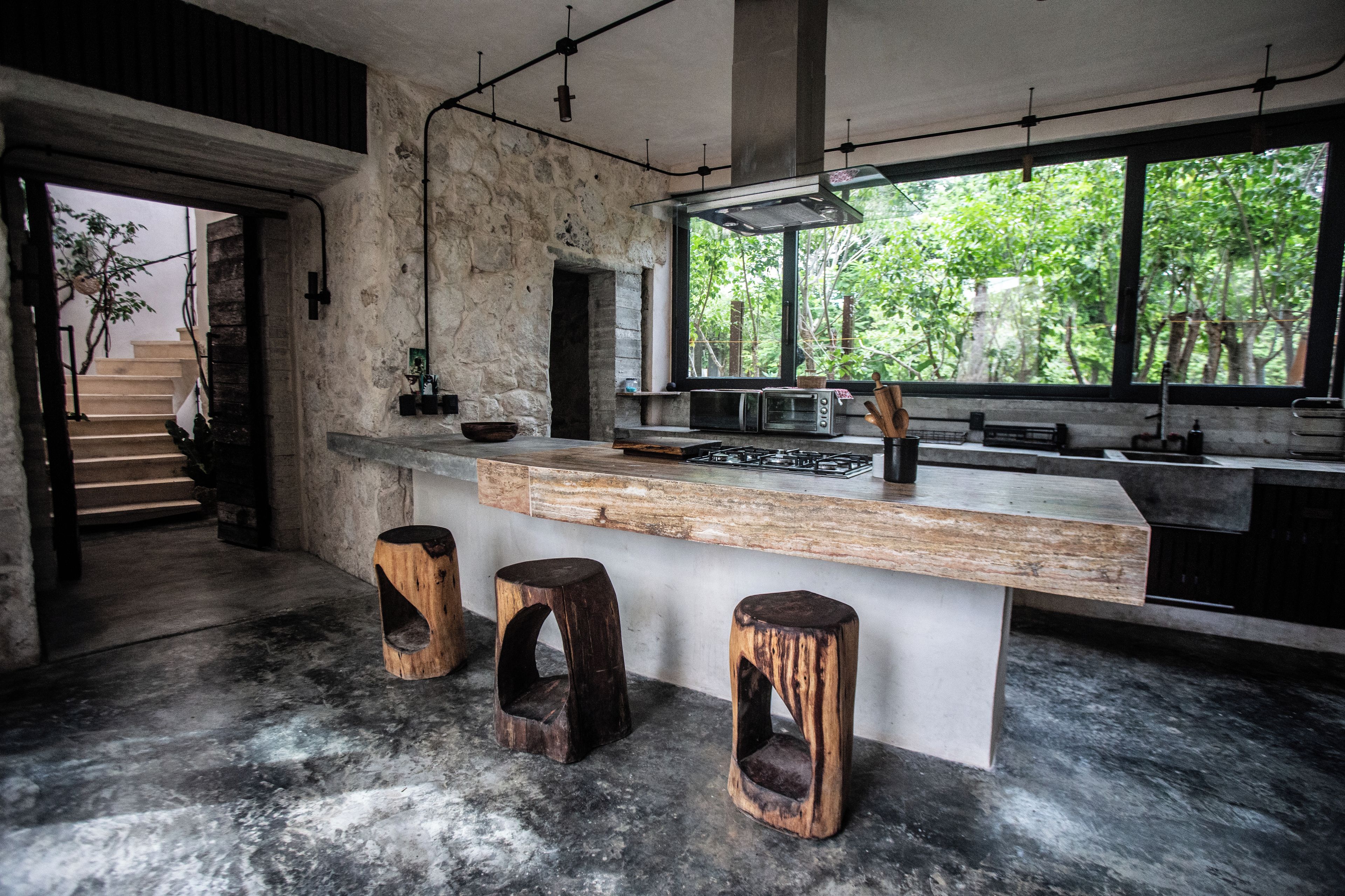 A modern kitchen with a large island, rustic stools, and a view of greenery outside.