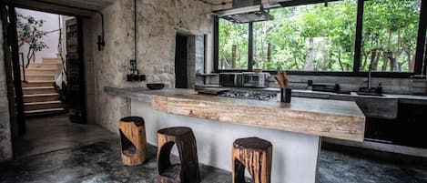 A modern kitchen with a large island, rustic stools, and a view of greenery outside.