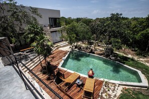 Une piscine avec une terrasse en bois, entourée d’arbres et bordée d’un bâtiment moderne.
