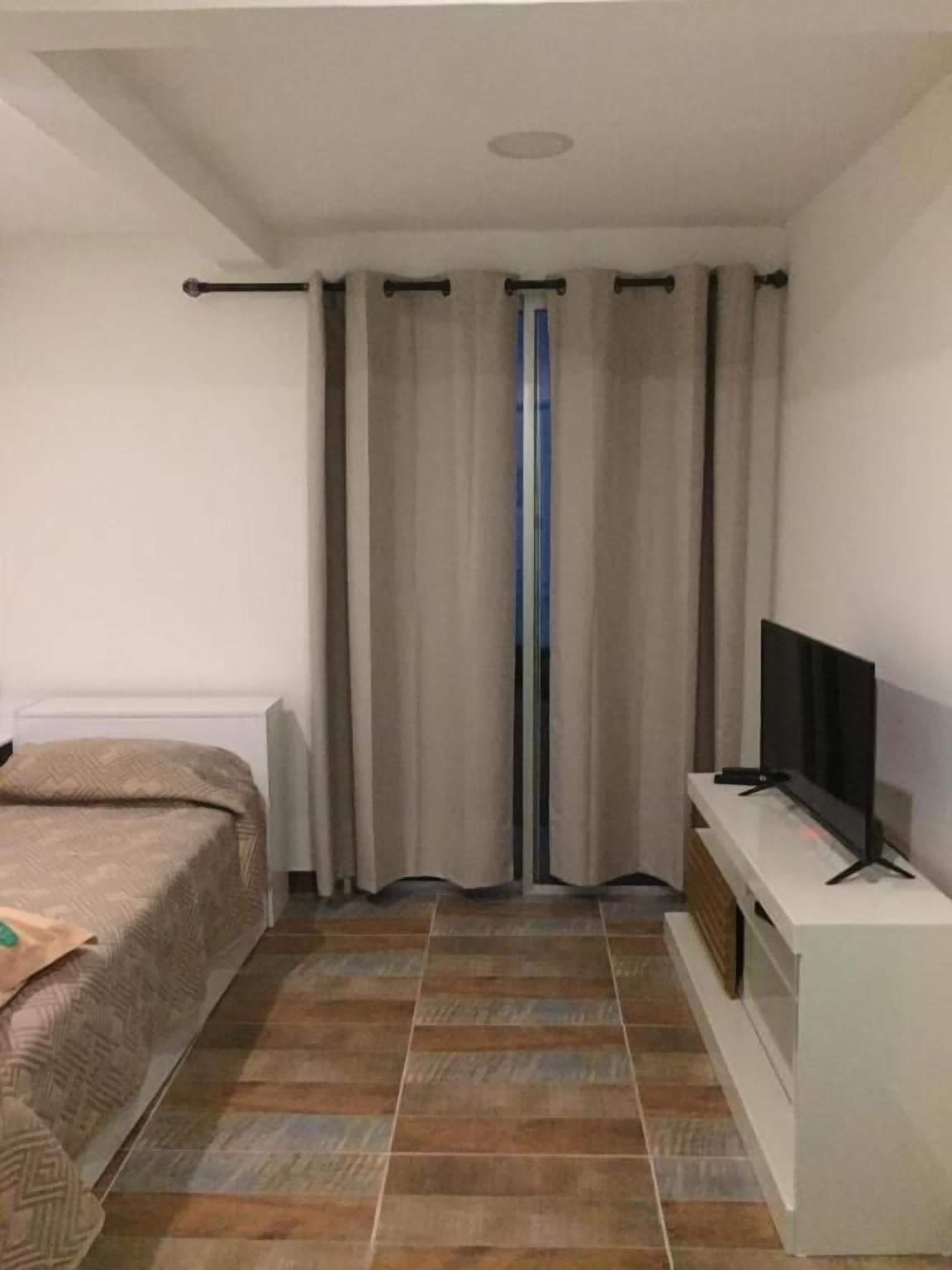 Une chambre à coucher avec un lit, un téléviseur posé sur un meuble, des rideaux et un sol carrelé.
