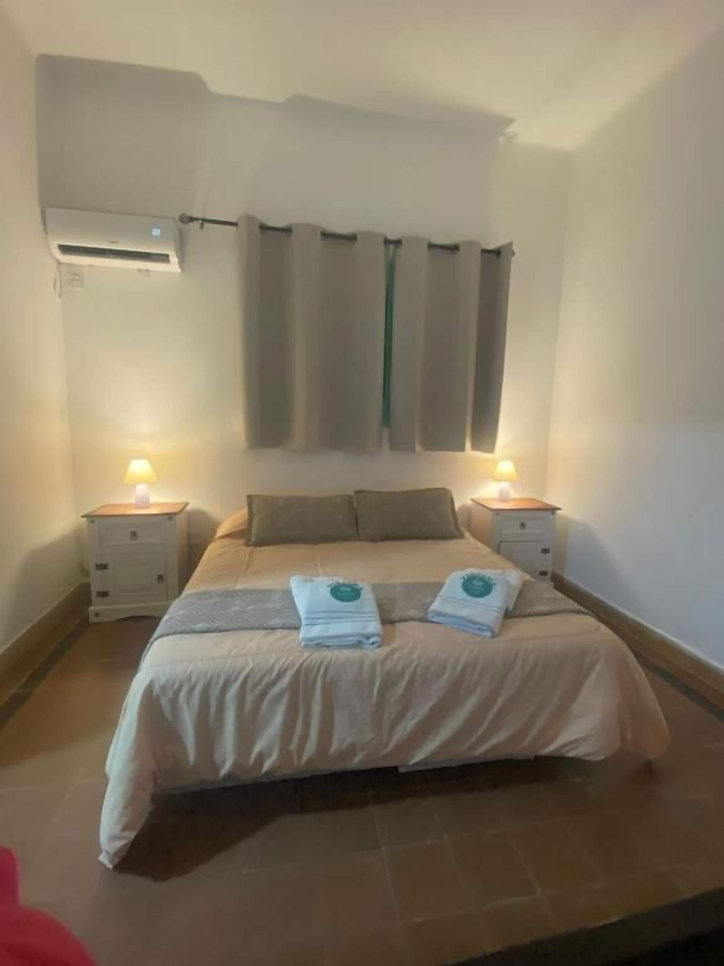 Un dormitorio con una cama, dos mesitas de noche, aire acondicionado en la pared y una cortina.